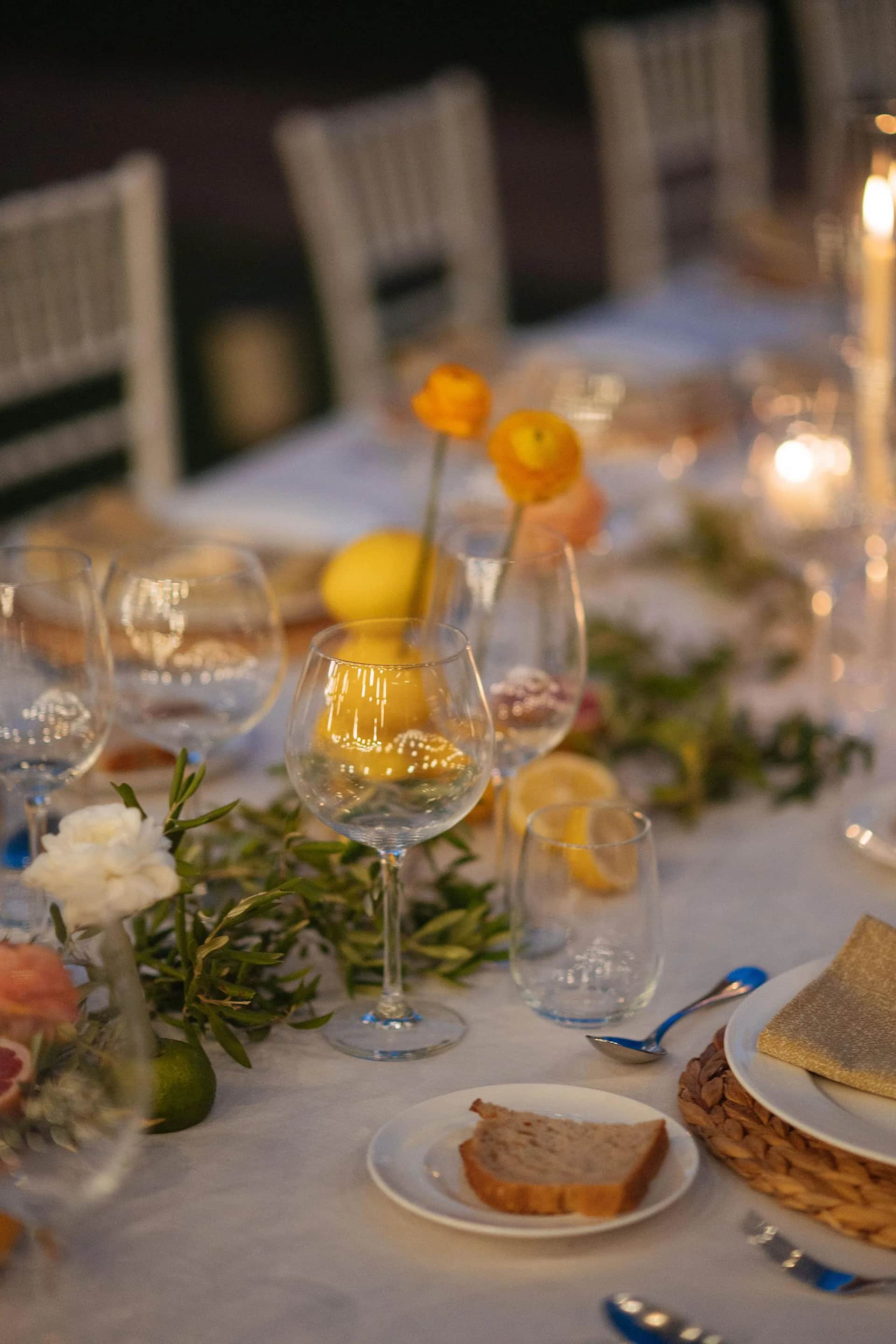 Détail d'arts de la table : verre à vin, pain, citrons et branches d'olivier sur nappe blanche