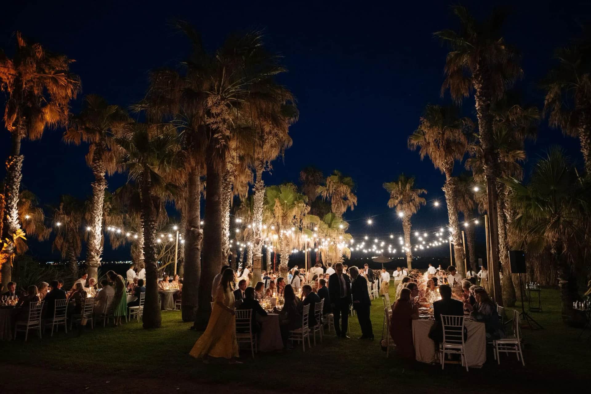 Dîner de mariage éclairé par des guirlandes guinguette tendues entre les palmiers, invités attablés