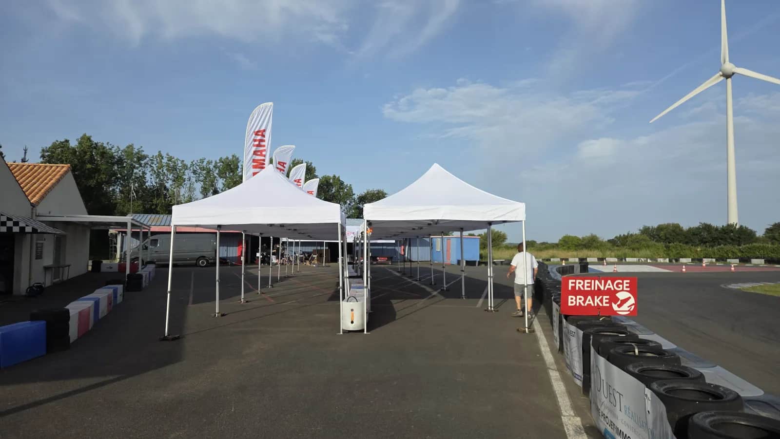 Pop-up gazebos set up near the paddock of the Mécamax circuit, "Freinage / Brake" sign and wind turbine in the background