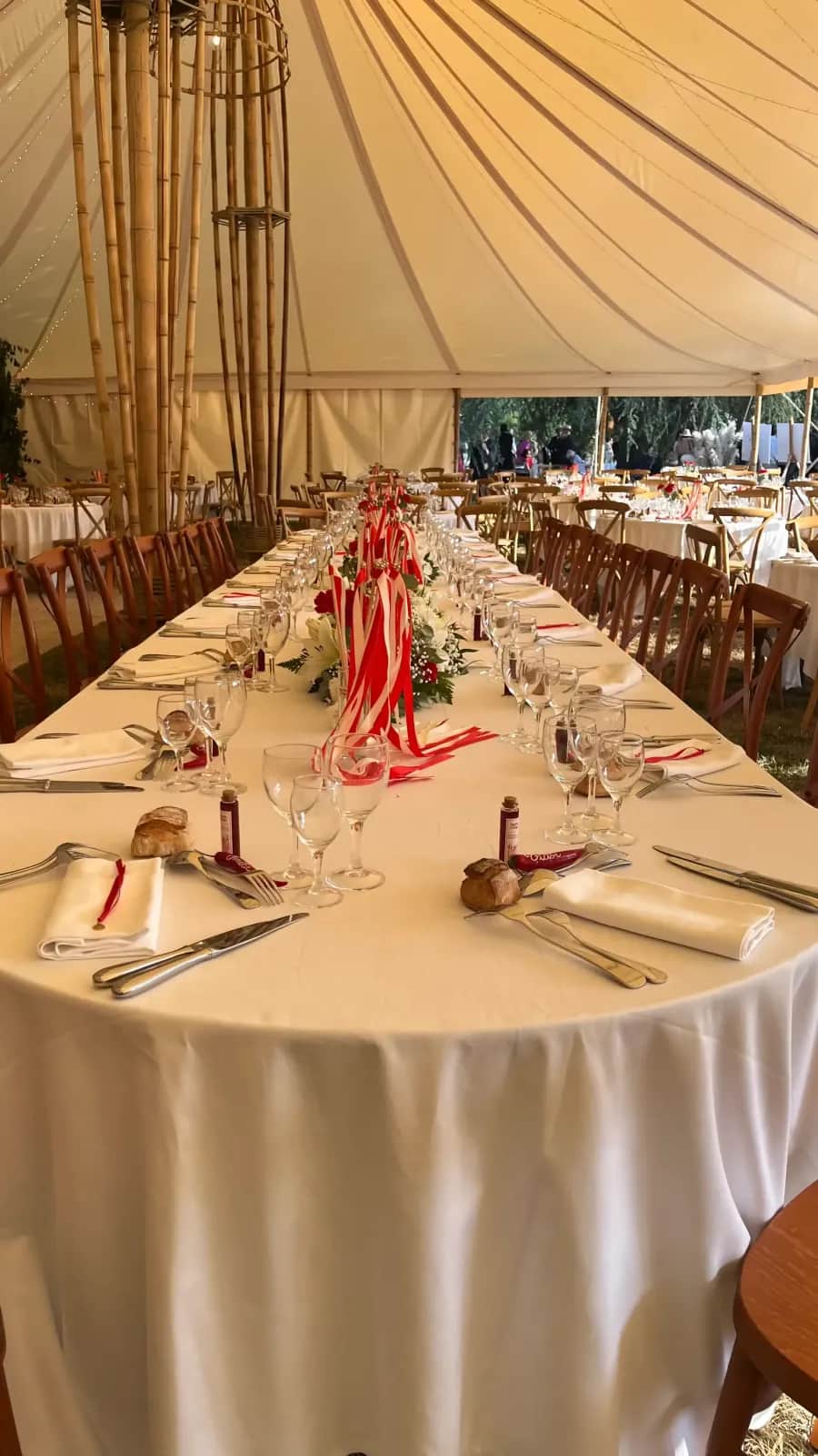 Wedding table runner: red and white ribbons, floral arrangements, glassware and bread on a long banquet table under the bamboo marquee